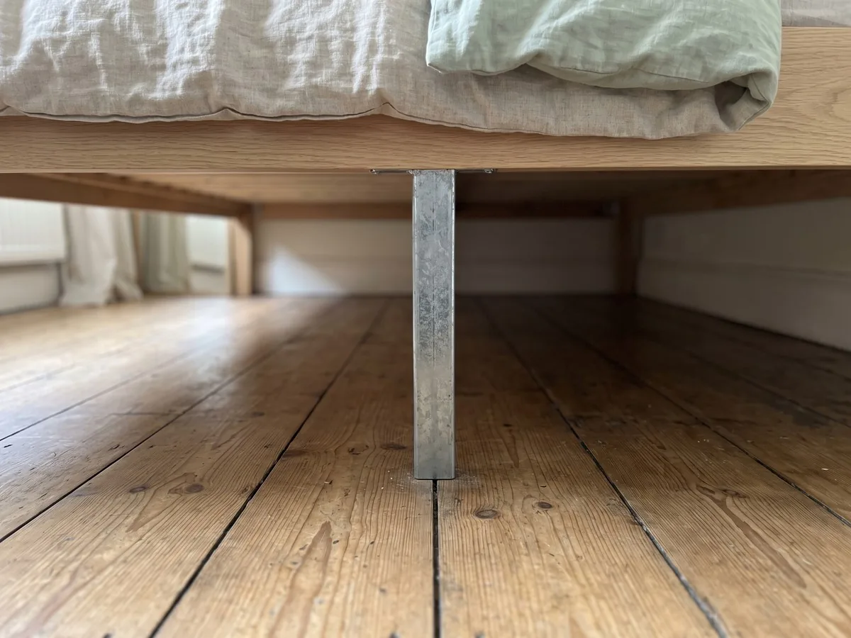 Floor-level view under a low pale-oak bed frame, where a galvanized steel center support leg stands on the floorboards in the middle of the space and meets a beam beneath the slats